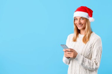Sarı saçlı, Noel Baba şapkası takan genç bayan akıllı telefonunu kullanmaktan zevk alırken gülümsüyor, parlak mavi arka planda neşeli ruhu somutlaştırıyor,
