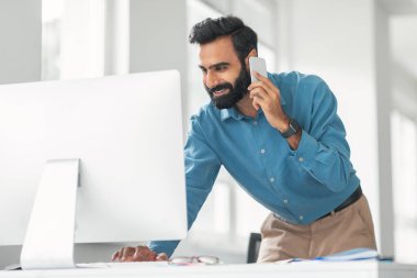 Mavi tişörtlü neşeli sakallı Hintli adam birden fazla iş yapıyor, telefonla konuşuyor ve bilgisayar ekranına bakıyor, çağdaş ofis içinde çalışıyor.