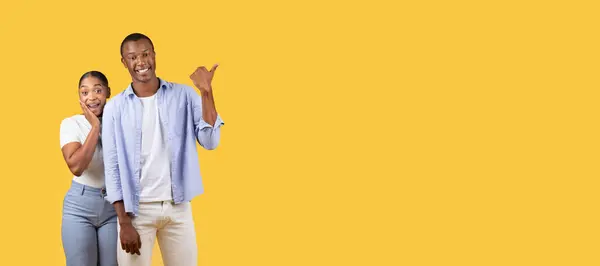 Cheerful black man pointing to the side while standing next to excited woman, both smiling with ample copy space on yellow background, panorama
