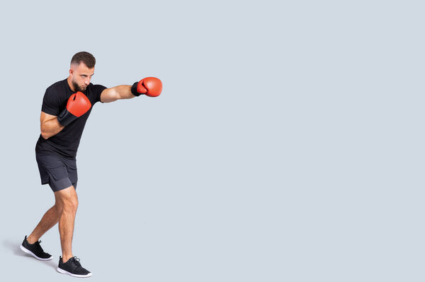 Serious millennial caucasian man in sportswear boxing gloves fight with fist, isolated on gray studio background, full length. Body care and professional workout, competition