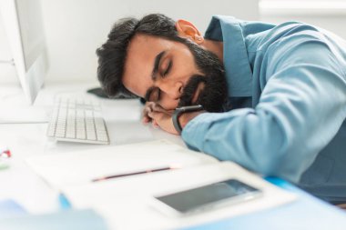 Beyaz ofis masasında kolunda sakallarıyla uyuyan yorgun Hintli işadamı, iş malzemeleriyle çevrili, kurumsal çevrede aşırı çalışma ya da yorgunluk taslayan