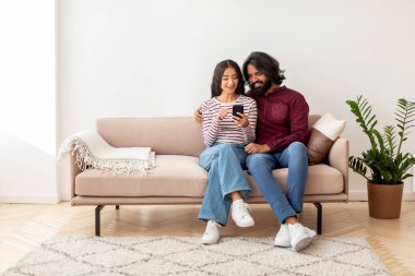 Olumlu sevgi dolu milenyum Hindistanlı çift güzel kadın ve erkek evde akıllı telefon kullanıyor, telefonla selfie çekiyor, ailesiyle görüntülü görüşme yapıyor, yeni mobilya seçiyor, internetten yemek sipariş ediyor, mobil uygulama kullanıyor.