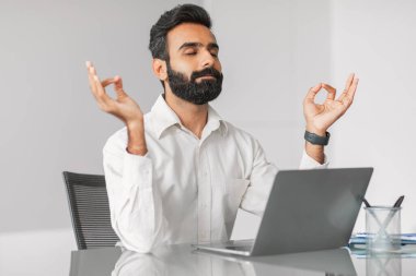 Ofis masasında meditasyon yapan beyaz gömlekli sakin Hintli işadamı, stresi azaltmak için, gözleri kapalı ve parmakları Mudra pozisyonunda, yoğun iş gününün ortasında.