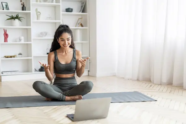 Young indian lady fitness coach yoga instructor have online class with student, sporty woman sitting on mat at home, looking at laptop screen, smiling and gesturing, copy space. Online workout