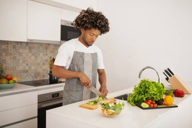 Önlüklü genç siyahi adam sağlıklı vejetaryen yemeği hazırlıyor, salata için sebze kesiyor, sağlıklı yaşam tarzını ve kilo kaybını somutlaştırıyor, modern mutfağın içinde duruyor.