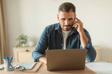 Yerel ofiste dizüstü bilgisayar kullanırken cep telefonuyla acil aramalara odaklanmış sakallı işadamı. Profesyonel iletişim, dijital iş akışlarının dengelenmesi