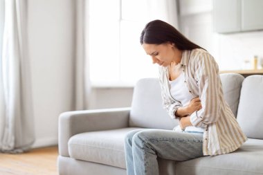 Sindirim sorunları. Akut karın ağrısından muzdarip genç bir kadının portresi, Avrupalı bir kadının regl sancısı ya da mide ağrısı, koltukta oturup karnına dokunması,