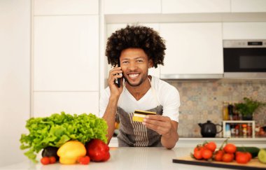 Genç Afrikalı Amerikalı bir adam kredi kartı tutuyor ve akıllı telefonunu yemek planlarını tartışmak için kullanıyor, online market alışverişi için ödeme yapıyor, modern mutfağında duruyor.
