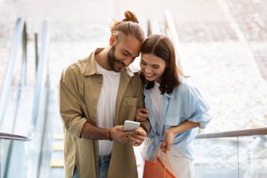 Gülümseyen beyaz kadın ve koca müşterileri alışveriş merkezlerinde akıllı telefonlara bakmaktan zevk alıyorlar. Uygulama ve satılık cihaz, alışveriş bağımlıları kazanır, nakde çevrimiçi sipariş