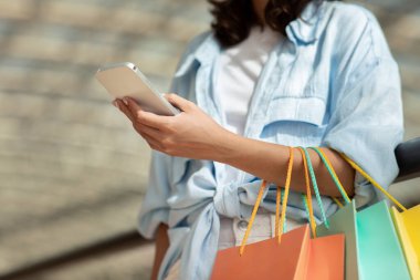 Elinde bir sürü alışveriş torbası olan Avrupalı genç bayan cep telefonuyla online alışverişin tadını çıkarıyor. Alışverişte kullanım uygulaması, reklam ve teklif, indirim, sipariş için ödeme, nakit ödeme