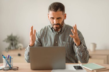Sakallı, orta yaşlı bir adam dizüstü bilgisayara kızgın, evde çalışma masasında oturuyor. İşadamı, dijital iş zorluklarını yansıtan bilgisayar sorunuyla karşı karşıya. Uzak görev sorunları