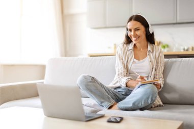 Kulaklık takmış genç bir kadın evinden dizüstü bilgisayarla internete giriyor, mutlu bir milenyum kadını bilgisayarda izliyor ve oturma odasında koltukta otururken not alıyor.
