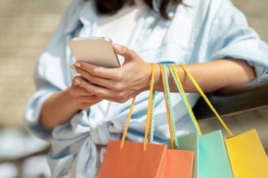 Elinde bir sürü alışveriş torbası olan bin yıllık beyaz kadın akıllı telefondan online alışverişin tadını çıkarıyor, kapatılıyor, kırpılıyor. Alışverişte yaşam tarzı, satış uygulaması, reklam ve teklif, indirim sezonu