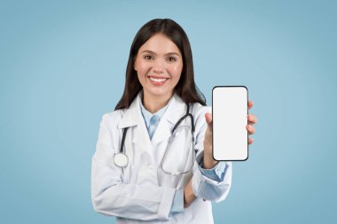 Neşeli genç bayan doktor boş akıllı telefon ekranı gösteriyor, steteskoplu laboratuvar önlüğü giyiyor, mavi arka planda dijital sağlık uygulamalarına hazır.