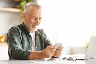 Mutfakta dizüstü bilgisayarla otururken mutlu yaşlı adam akıllı telefon kullanıyor, cep telefonuna gülümsüyor ya da yeni uygulamalara bakıyor, fotokopi uzayı ile yakın çekim yapıyor.