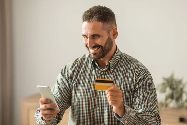 Middle aged man smiling while shopping and making payments online with his cellphone and credit card, sitting at domestic office, exploring internet bank offers in mobile application