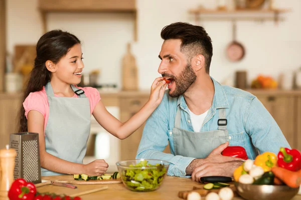 In warm home setting, a cheerful young girl gives slice of fresh tomato to her delighted father during fun and interactive healthy cooking session