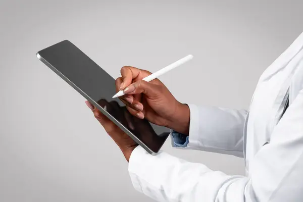 Black millennial lady doctor in white coat write on tablet screen, isolated on gray background studio, close up. Health care recipe online, medical help with device app, prescription