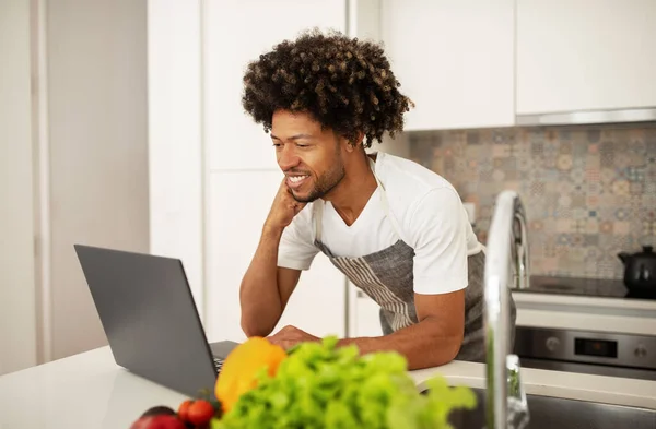 Smiling black man in apron learning to cook via laptop, prepping a healthy meal with fresh groceries products and websurfing on computer at kitchen indoors. Tech meets nutrition