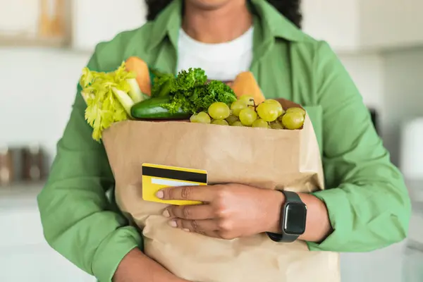 Cropped shot of African American woman holding her credit card and delivered grocery order in paper shopping bag, posing in modern kitchen interior. Unrecognizable buyer with food products