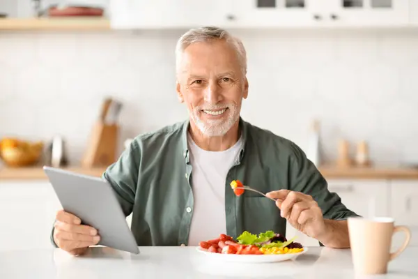 Happy Older Man Having Breakfast And Using Digital Tablet In Kitchen, Smiling Senior Gentleman Reading News, Browsing New App Or Shopping Online With Modern Gadget At Home, Closeup