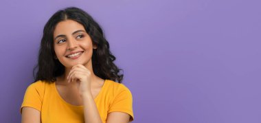 Heyecanlı, kıvırcık, sarı tişört giyen, çenesine dokunan, mor renkli stüdyo arka planında izole edilmiş reklam için fotokopi odasına bakan,