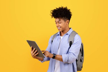Siyahi adam dijital tablet kullanan, sırt çantası takan, sarı stüdyo geçmişine karşı duran, modern eğitimi somutlaştıran bir öğrenci.