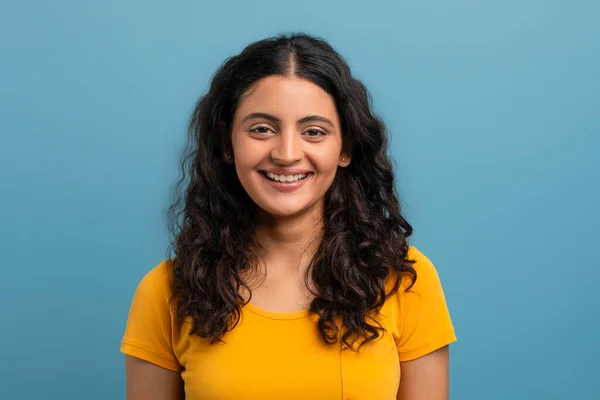 カメラで笑顔でブルーカラースタジオの背景に隔離された黄色のTシャツを着ているかなりキュリーな若いインドの女性学生 — ストック写真