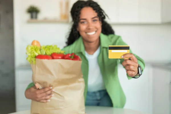 Cheerful black woman holds credit card and paper bag with grocery products at modern kitchen, celebrates convenience of online groceries shopping and joy of healthy nutrition. Selective focus
