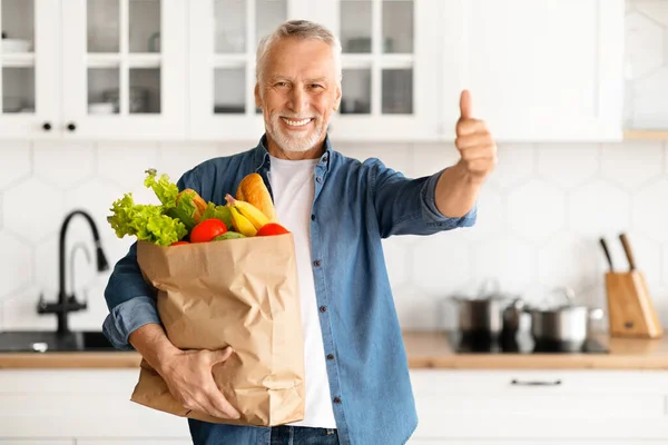 Happy Elderly Man Holding Paper Bag With Groceries And Showing Thumb Up, Cheerful Senior Gentleman Carrying Purchases And Smiling At Camera While Standing In Kitchen And Home, Free Space