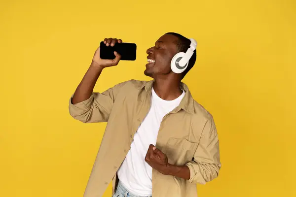 Cheerful young black guy in casual wireless headphones sing at mobile smartphone with empty screen, enjoy spare time, fun, isolated on yellow studio background. Ad and offer, app for karaoke