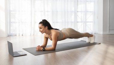 Formda ve kahverengi saçlı güzel bir kadın evindeki yoga minderinde esneme hareketleri yapıyor. Bilgisayarındaki egzersiz videosunu izliyor. Mesaj için boş bir alan, reklam.