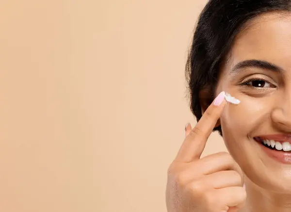 Half Face Portrait Of Young Indian Woman Applying Under Eye Cream And Smiling At Camera, Happy Beautiful Eastern Lady Moisturising Skin, Posing Over Beige Studio Background, Copy Space
