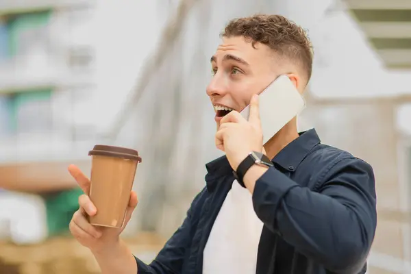 Glad surprised young caucasian man enjoy cup of coffee, calling by mobile phone, got good news, talk, point finger at free space in city. Work business ad and offer, communication surprise emotions