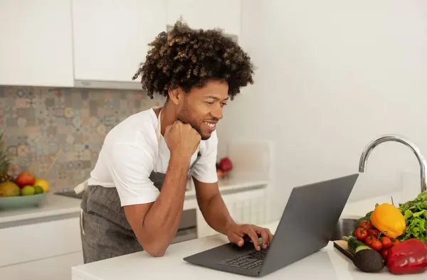 African American guy browsing recipes online on laptop computer, preparing vegetables for dinner in modern kitchen, merging culinary skills with digital e-learning. Cooking website ad
