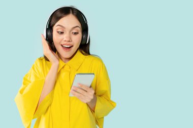 Müzikten hoşlanan, podcast dinleyen, yeni kablosuz kulaklık ve akıllı telefon kullanan, müzikal mobil uygulamaları kontrol eden, kopyalama alanı olan, mavi stüdyo arka planında izole edilmiş mutlu genç bir kadın.