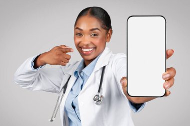 Beyaz önlüklü siyahi kadın doktorun, gri arka plan stüdyosunda izole edilmiş beyaz ekranlı akıllı telefona parmak atmasına sevindim. Sağlık, reklam ve teklif, uygulama tavsiyesi, profesyonel danışmanlık