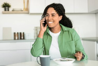 Gülümseyen Afrikalı Amerikalı kadın, modern mutfakta kahvaltı ederken akıllı telefonlardan konuşuyor, masada oturuyor ve telefon görüşmeleri sırasında sağlıklı mısır gevreklerinin tadını çıkarıyor.