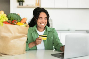 Evde modern mutfak masasında oturan, alışveriş ürünleri satan ve web sörfü yapan kredi kartı ile gülümseyen genç siyahi bir kadın.