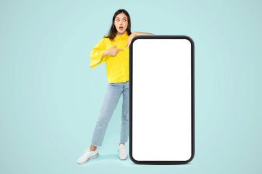 Şaşırtıcı derecede şık bir milenyum kadını mavi stüdyo arka planında beyaz ekranlı büyük bir telefonu işaret ediyor. Model, reklam için fotokopi alanı, mobil uygulama, online teklif.