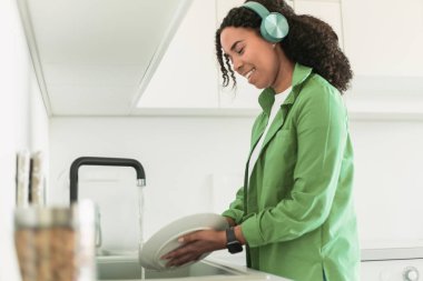 Kulaklıklı neşeli siyahi bayan mutfaktaki lavaboyu temizlerken, bulaşıkları yıkarken ve günlük işleri boş zamanlarla harmanlarken müzik veya sesli kitap dinliyor. Yan görünüm
