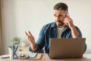 Memnun olmayan sakallı, orta yaşlı bir adam, rahatsız edici sorunlu telefonlara cevap verirken bilgisayarını karıştırıyor, serbest çalışanların yoğun yaşamlarını ve iş sorunlarını özetliyor, modern ev ofisinde oturuyor.