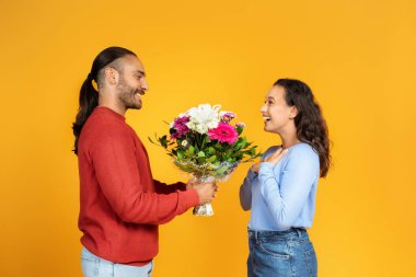 Pozitif milenyum çocuğu sarı stüdyo arka planında izole edilmiş bayana bir buket çiçek veriyor. Doğum günü tebriği, yıldönümü sürprizi, birlikte bayram kutlaması ve romantik randevu.