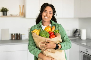Afro-Amerikalı bir kadın mutfak içinde alışveriş poşetlerine sarılıyor. Tatmin olmuş bir alıcı marketten ürünleri getirdi, kameraya gülümsedi, sağlıklı beslenmeyi destekledi.
