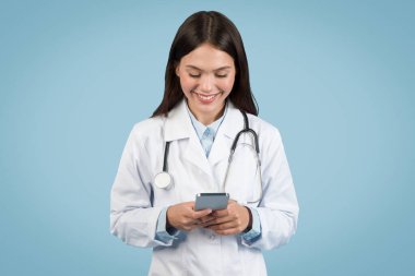 Steteskoplu laboratuvar önlüklü neşeli kadın doktor, temiz, açık mavi arka planda hastayla mesajlaşarak akıllı telefonuna odaklanmış durumda.