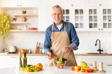 Yemek pişirme konsepti. Mutfakta taze sebze salatası hazırlayan mutlu kıdemli adamın portresi kasede önlük takan yaşlı beyefendinin gülümsemesi evde lezzetli yemekler yapması, uzay kopyalaması.