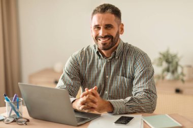 Kafkasyalı orta yaşlı bir adam dizüstü bilgisayarla çalışırken gülümsüyor, modern dahili ofis içinde çalışma masasında poz veriyor, kariyerini internet üzerinden yönetiyor. Başarılı uzaktan çalışma ve serbest çalışma