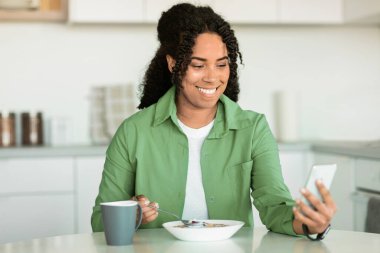 Gülümseyen Afrikalı Amerikalı kadın mısır gevreğini, kahvaltıyı ve kahveyi, internetteki tarifleri, mobil uygulamaları ve sosyal medyada mesajlaşmayı sevdiği için akıllı telefon kullanıyor.