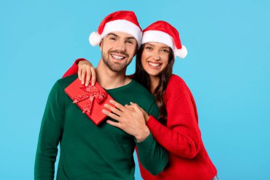 Romantik Noel Hediyesi. Noel Baba şapkalı mutlu çiftin portresi Xmas hediye kutusuyla poz veriyor, kocasına sarılan kadın kış tatilinde mavi stüdyo arka planında hediyesiyle ona sürpriz yapıyor.