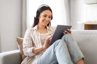 Ev eğlencesi. Evdeki koltukta dijital tabletle kablosuz kulaklık takan, gülümseyen, sosyal ağlara bakan, modern Gadget, yakın plan